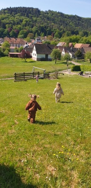Zwei Kinder laufen über eine Wiese, im Hintergrund das Dorf und der Wald